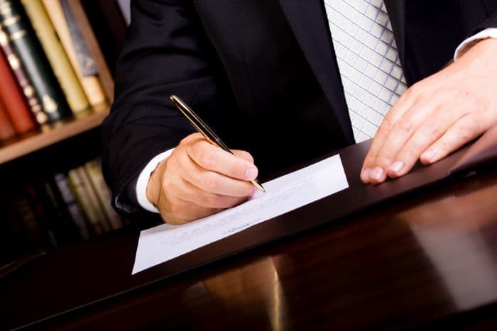 Person wearing a black suit with silver tie writing with a 400TN gold titanium nitride bullet space pen on a paper on a wooden surface. There are books on a bookcase in the background.